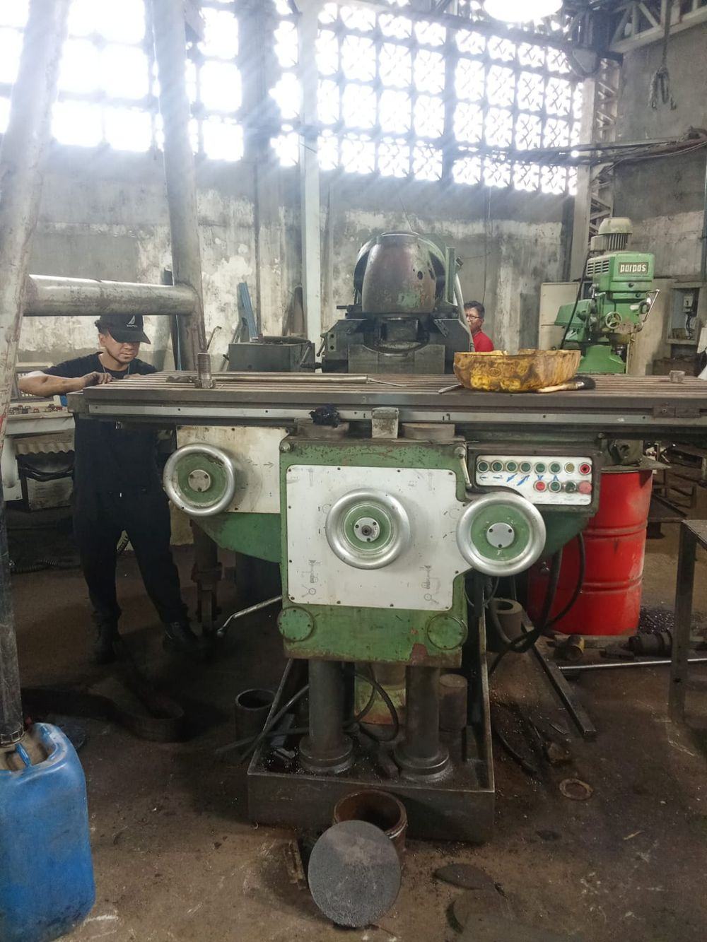 Fresadora industrial verde con mesa de ranuras en T, panel de pulsadores y tres volantes de mando, con un operario junto a la máquina